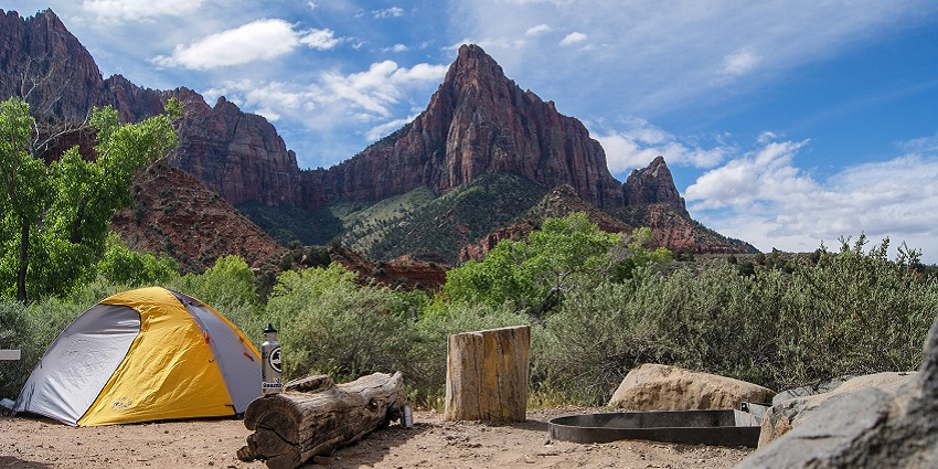Between the mountains, a tent is placed near a log of wood and a bottle of water