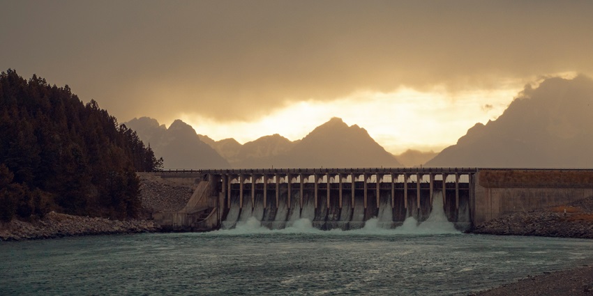 Sunset view of a dam with gushing water, surrounded by pine trees and mountain peaks.