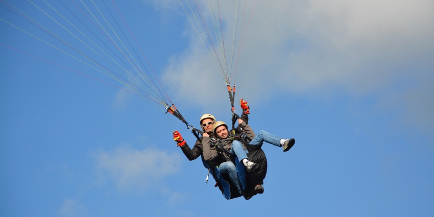 Two people tandem paragliding in a clear blue sky with the pilot controlling the fly
