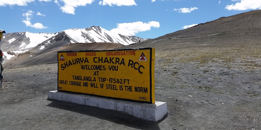 Board of the Tanglang la peak on a rocky plain with snow covered peaks far behind