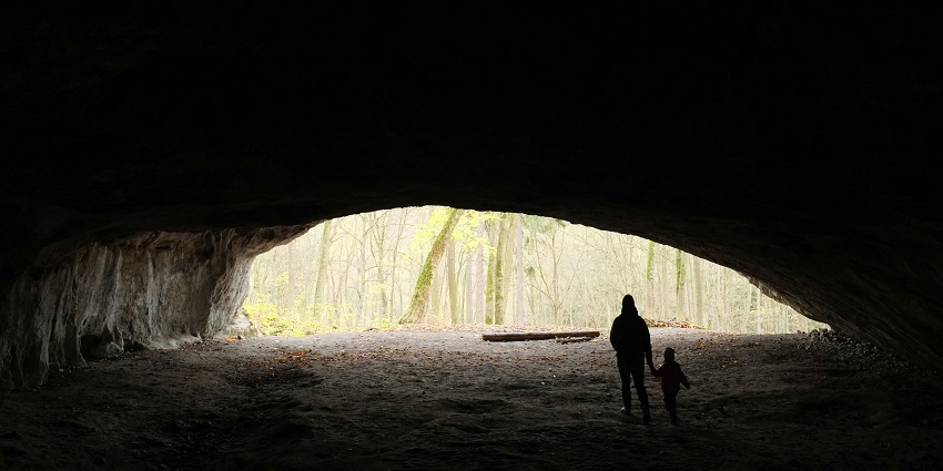 A person with a child is standing inside a big-mouthed cave, holding each other’s hands.