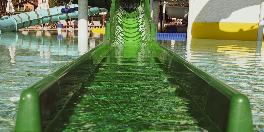 View of a water slide leading to a pool at an amusement park, surrounded by green trees.