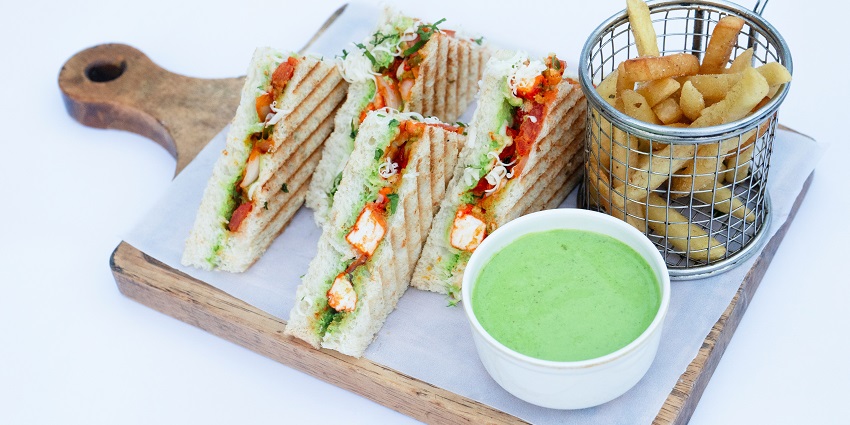 An image of a tray filled with four veg sandwiches, french fries and a chilli sauce.
