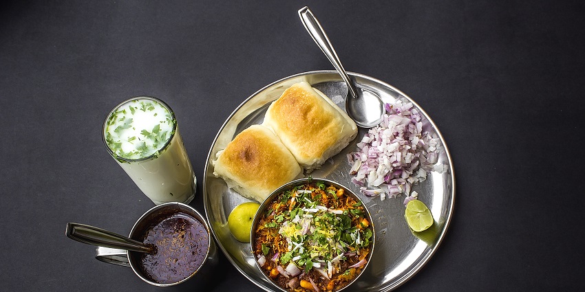 Image of a plate of misal pav served with onions, lemons, red chutney and a glass of lassi