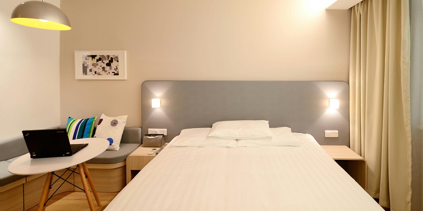 A room with fluorescent lighting and a clean bed with a laptop placed on the side table