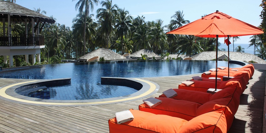 Poolside view of a lodge with seating area, an amazing ambience, and many coconut trees around it.