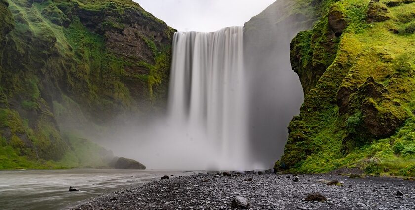 Zenith waterfall cascading down a rocky cliff into a misty river, surrounded by lush greenery