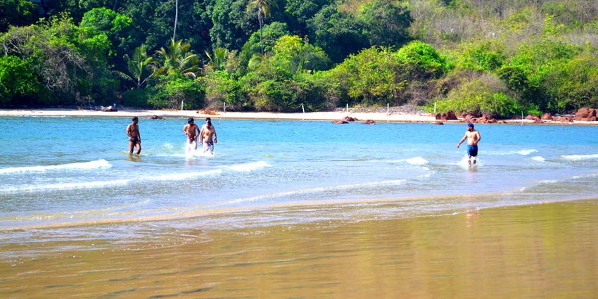 Beach with clear blue water and gentle waves, surrounded by nice greenery and rocky cliffs.