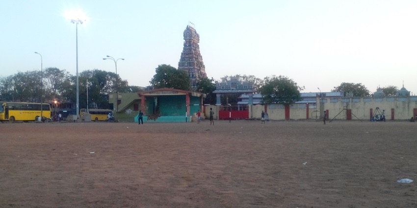 The tower of a temple is shown with few people standing outside and two buses parked.
