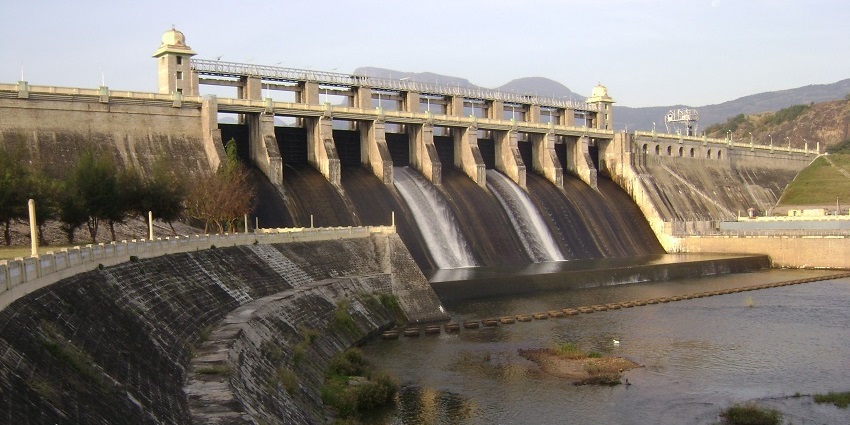 Amaravathi Dam with water flowing through spillways, surrounded by hills and greenery