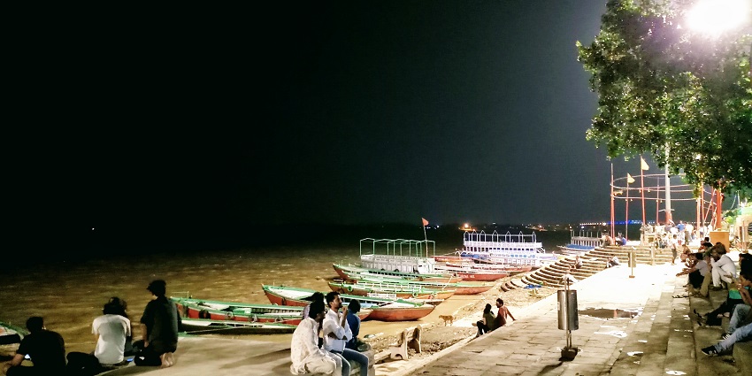 A vibrant scene of Assi Ghat in Varanasi at sunset, with people gathered by the riverbank.