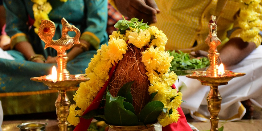 Two people engaging in a pooja ritual with a dry coconut covered in garland and leaves