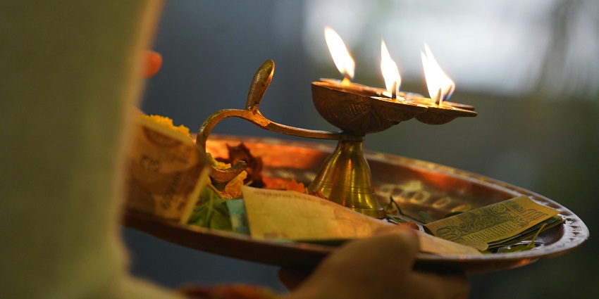 A person holding a brass puja tray with a burning diya, money, flowers and leaves.