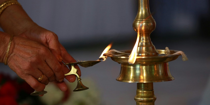 A person lighting diyas from a tall brass diya and glowing in a dimly lit setting.