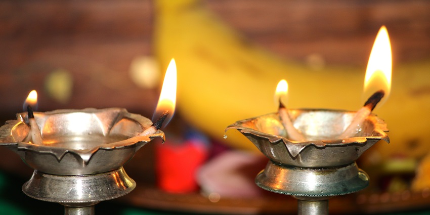 Two brass diyas burning on an alter with a banana on a tray in a blurred background.