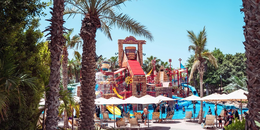 People are walking by a swimming pool surrounded by palm trees and lounge chairs on a sunny day.