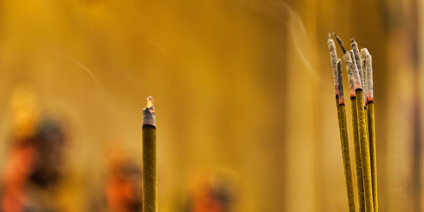 Burning incense sticks emitting smoke, placed in a sandy holder with a blurred background