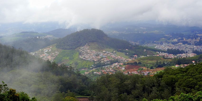 An aerial view of Ooty, showcasing hills, winding roads, and scattered houses under a clear sky.