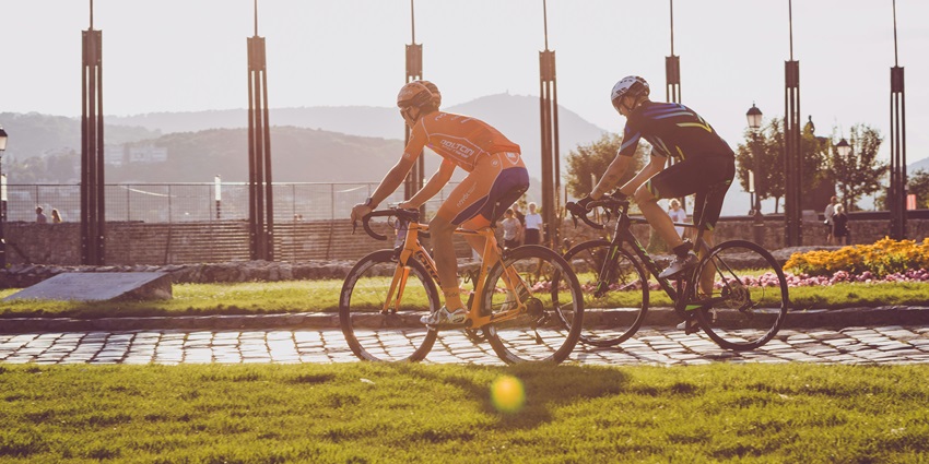Two cyclists ride along a path during sunset, with flags and hills in the background.