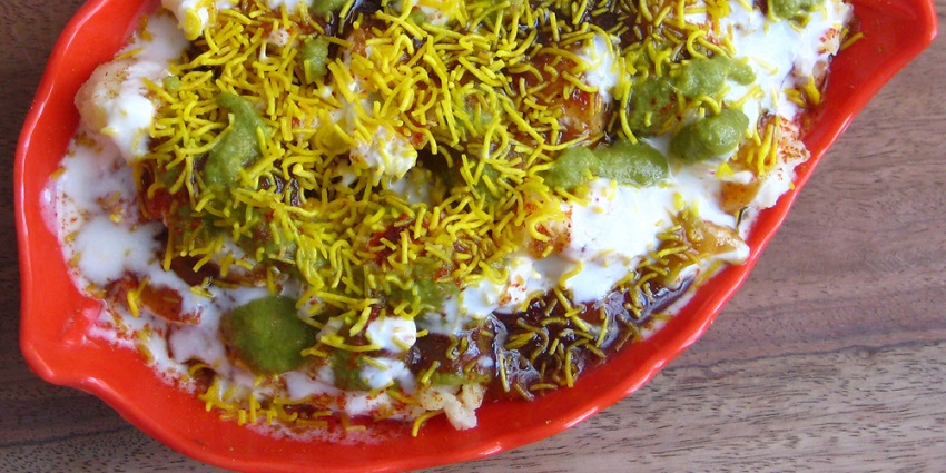 Papdi chaat is served in a red bowl topped with a layer of curd and green sauce