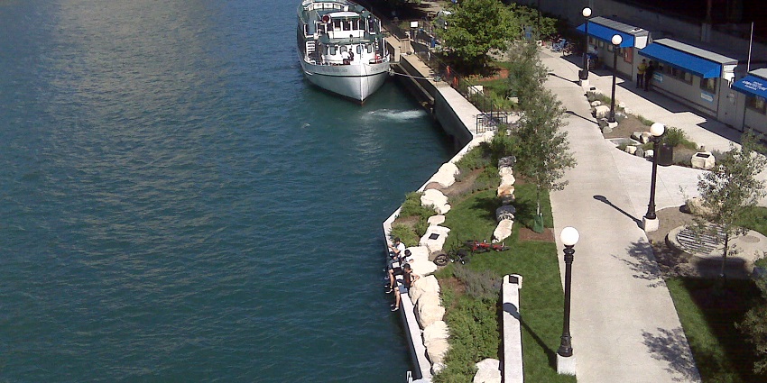 Boats parked at the Chicago Riverwalk clean water with lamps and trees on the pathway