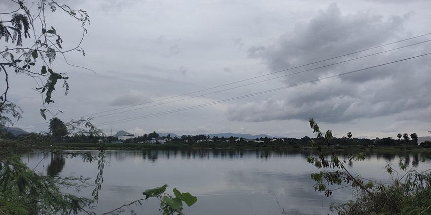 Serene lake is surrounded by lush greenery, reflecting the clouds above and distant hills.