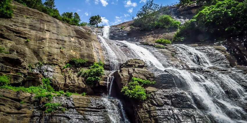 Water flows off rock cliffs in the form of a fall against a clear blue sky and a couple of trees.