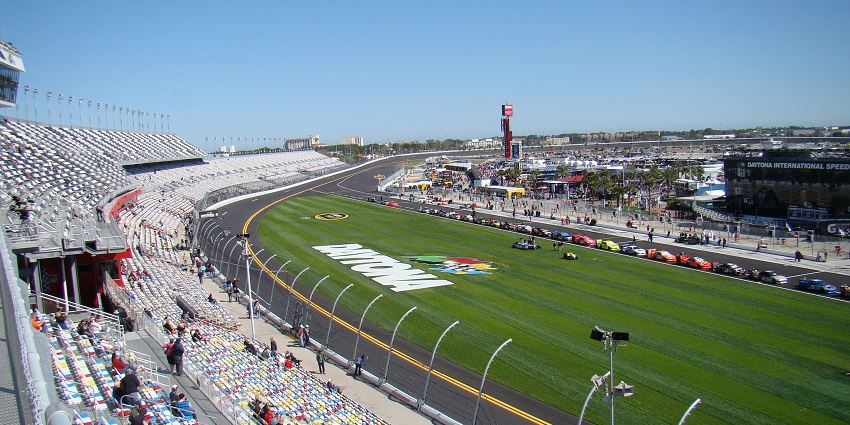 Daytona International Speedway with sports cars parked and Daytona painted on the grass.