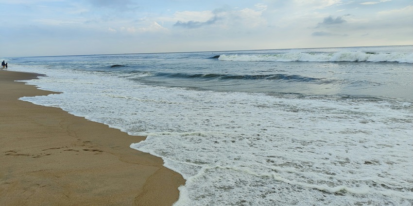 The calm Besant Nagar, Chennai, with gentle waves, a cloudy sky, and footprints in the sand.