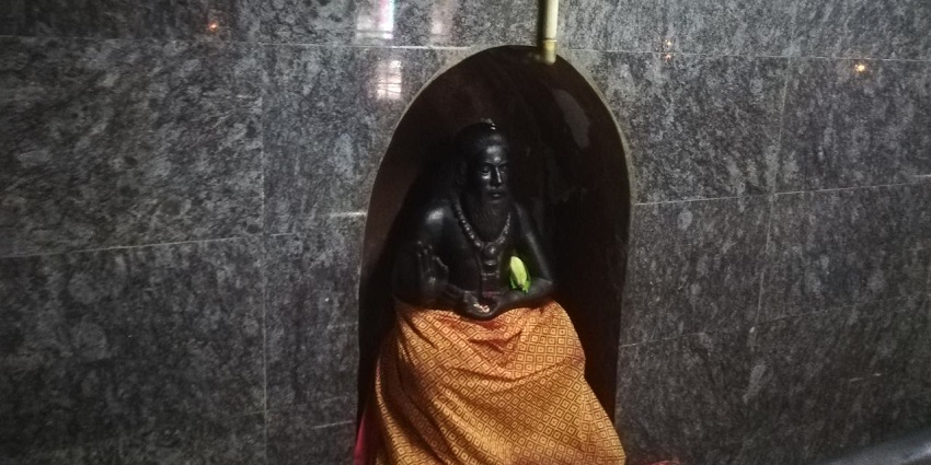 A glimpse of Thiruvalluar idol decorated with clothes sitting at the Putlur temple under a sign.