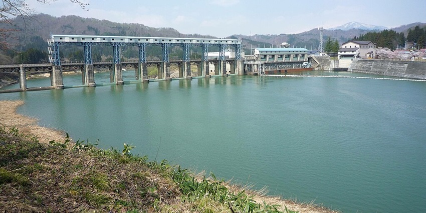 Large dam structure with flowing water, surrounded by lush greenery and a clear blue sky