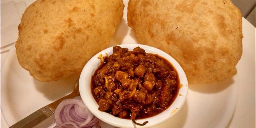 Two pieces of bhatura are placed on a plate alongside chola and onion slices on the side