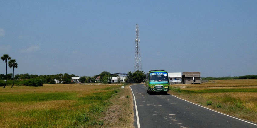 A bus travelling on a rural road in Tamil Nadu, surrounded by green fields and trees to reach Thiruvaiyaru temple.