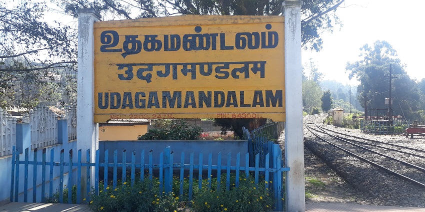 A photo of Ooty railway station with red-roofed buildings, blue signboard, and green hills.