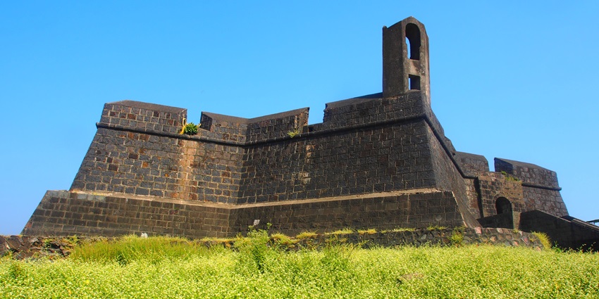An ancient fort, characterised by its sturdy stone walls and arched gateways, surrounded by palm trees and lush greenery, with a backdrop of a clear blue sky.