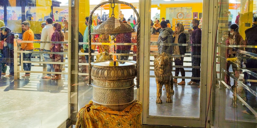 A Jyoti lamp placed in a glass chamber with people waiting in a queue outside the temple