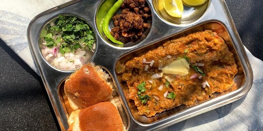 In a utensil Pav bhaji is served accompanied by lemon wedges and shredded onions