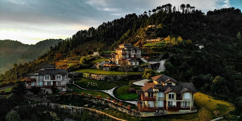 A white and brown hilltop house overlooking terraced green slopes, set in a serene mountain environment in Kanatal, one of the best places to visit in Uttarakhand in October.