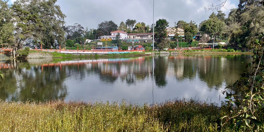 Serene Kodaikanal Lake reflects trees and mountains, with calm waters and distant houses