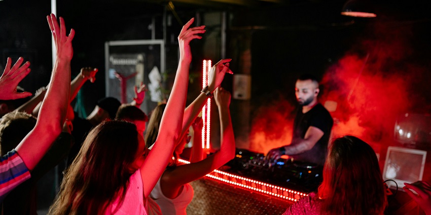 People dancing inside the club with lots of lights, lasers, a DJ and an LED screen.