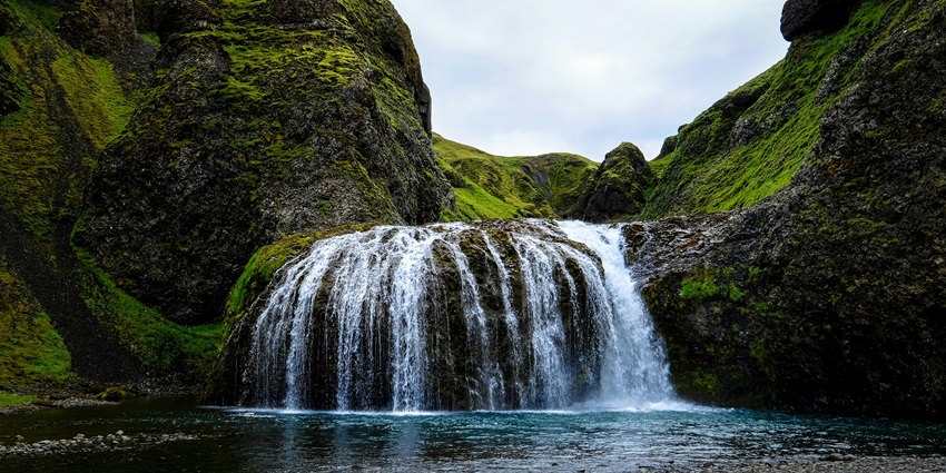 Image of a waterfall flowing down rocky cliffs surrounded by greenery under a cloudy sky.