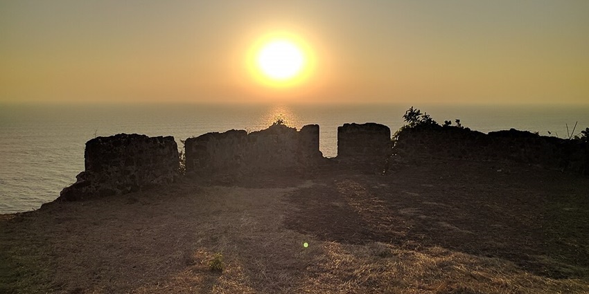 A silhouette of a ruined Korlai Fort against a vibrant sunset over the ocean.