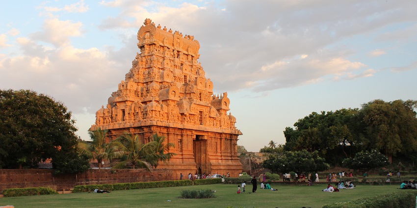 People sitting on the grounds near the Brihadiswara temple surrounded by greenery.