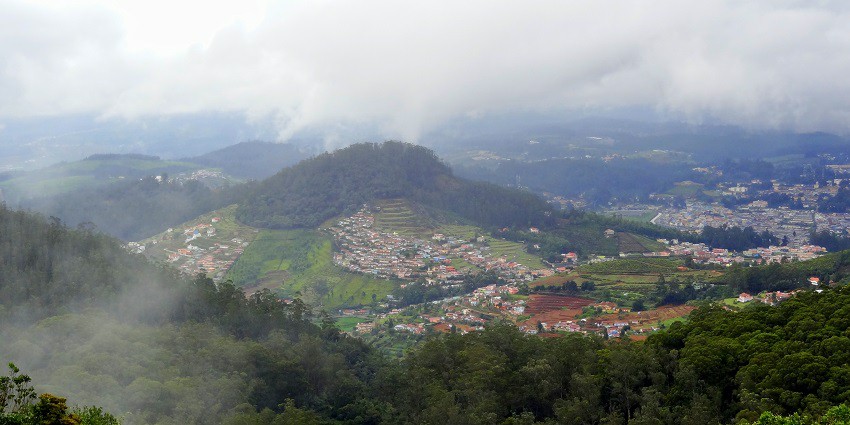 An aerial view of Ooty town, showcasing buildings, greenery, and surrounding hills under a cloudy sky.