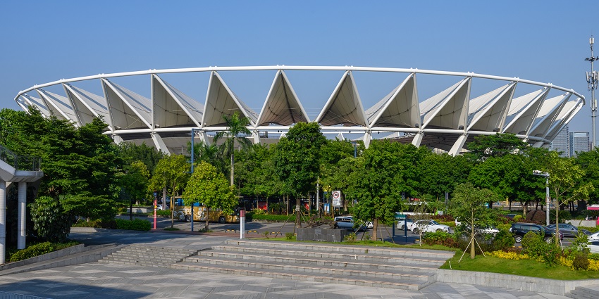 Image of stadium from the outside with walkway surrounded by lush green tree and cars parked near the parking area
