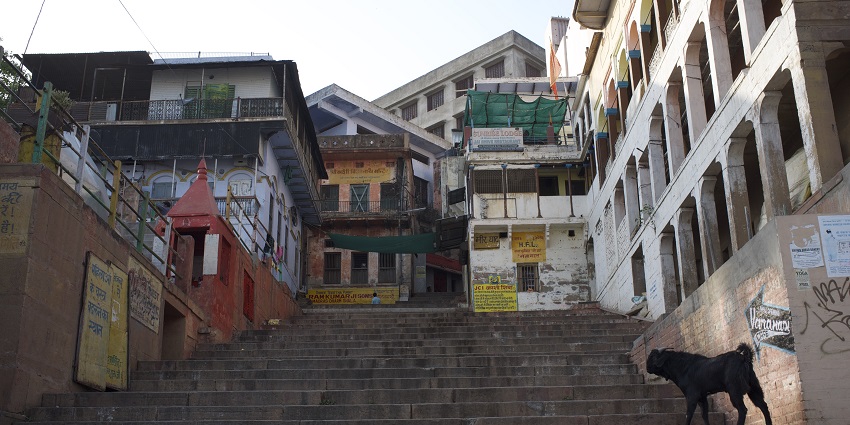 A serene view of Varanasi Street surrounded by buildings and boats lines the Ganges River.