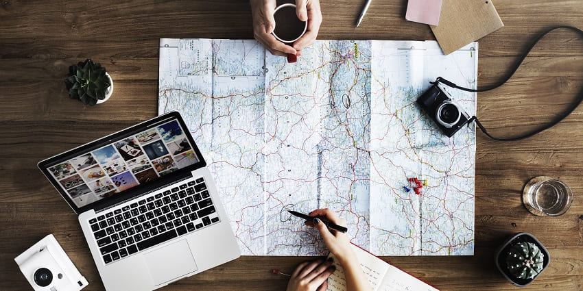 Image showcasing a map, laptop, dairy and pen symbolising planning for a trip over coffee.