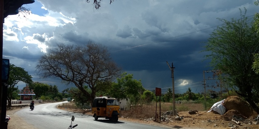 Road with a motorcycle, auto-rickshaw passing through and cloudy sky suggesting rain