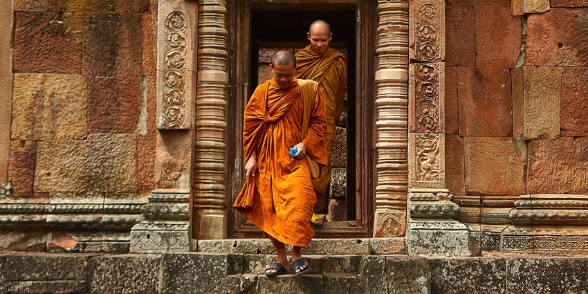 Two monks dressed in traditional orange robes walk down a monastery with green trees.