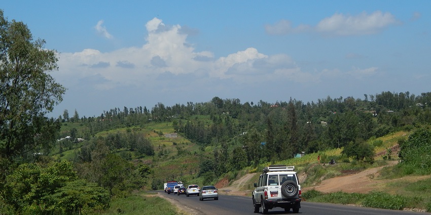 A hillside road with multiple cars surrounded by greenery and distant houses on hills.